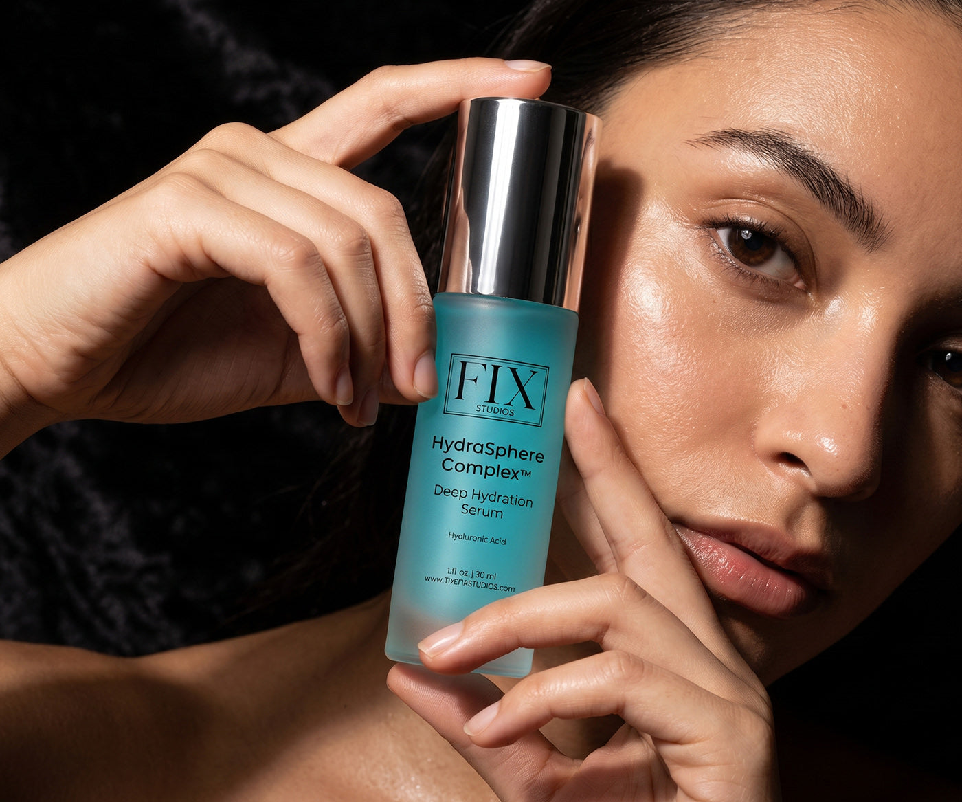A woman holds near her face a bottle of Fix Studios HydraSphere Complex Hyaluronic Acid Deep Hydration Serum for deeply moisturized skin and a protective barrier that lasts all day