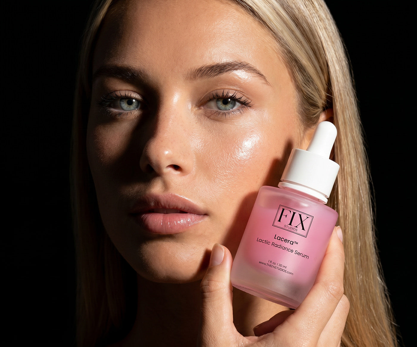 A beautiful woman holds a bottle of Fix Studios medical-grade Lacera™ Lactic Radiance Serum close to her face on a black background.